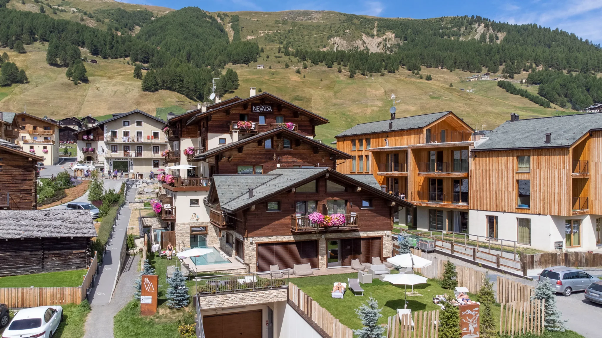 Vista aerea dell'hotel a Livigno con chalet in legno, giardino fiorito e montagne verdi sullo sfondo
