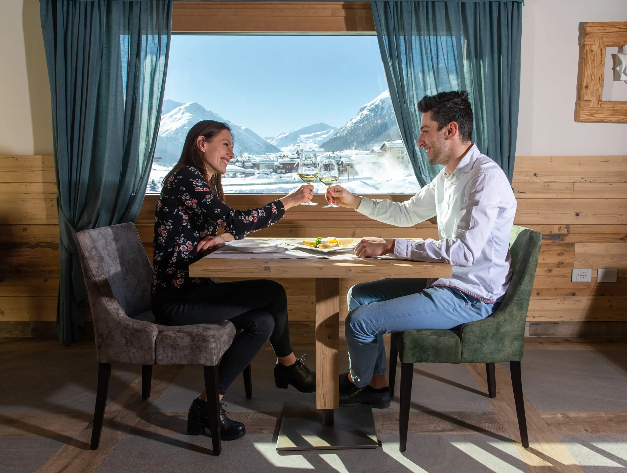 Coppia brinda con calici di vino a tavola davanti a una finestra con vista sulle montagne di Livigno