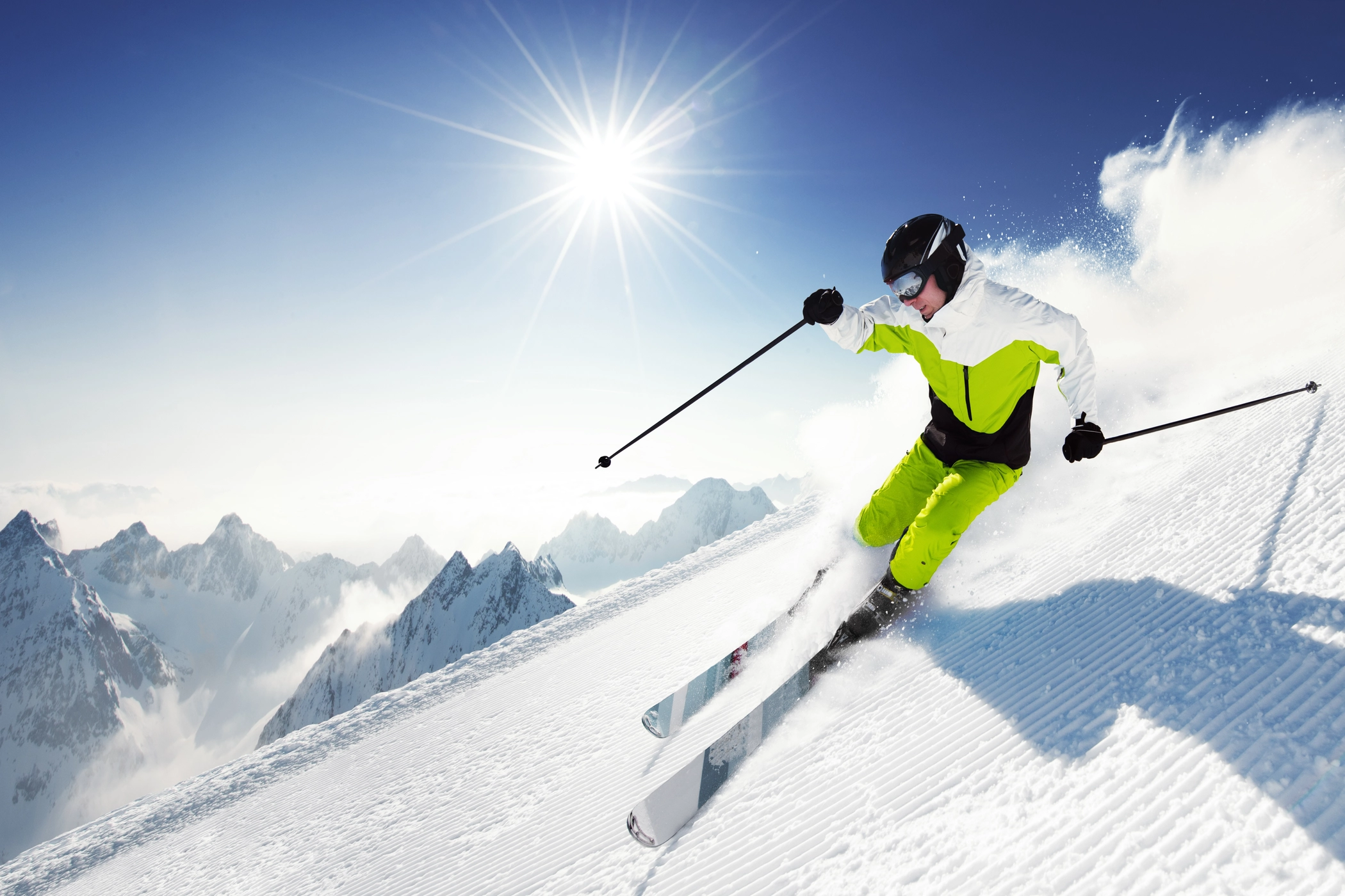 Sciatore in tuta verde scende su pista innevata con montagne e sole splendente sullo sfondo