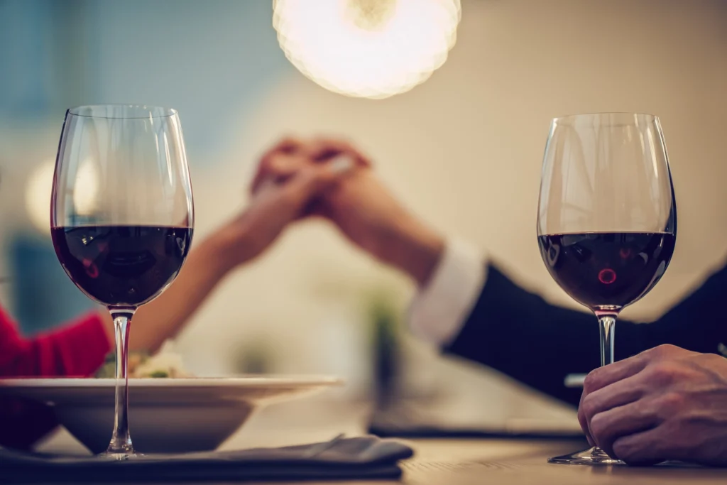 Coppia si tiene per mano a cena romantica con due calici di vino rosso in primo piano